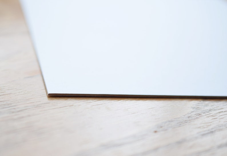 May include: A close-up of the edge of a white sheet of paper on a light brown wooden surface.