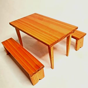 Peut inclure: Un ensemble de table et de deux bancs en bois. La table et les bancs sont en bois brun clair avec une finition naturelle. La table est rectangulaire et a quatre pieds. Les bancs sont également rectangulaires et ont quatre pieds.