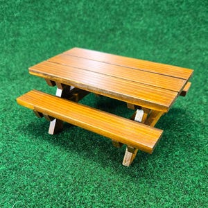 May include: A miniature wooden picnic table with a warm brown finish. The table features a slatted tabletop and attached bench, perfect for small-scale displays or dollhouse settings. The table is set against a green background.