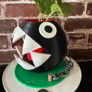 Może przedstawiać: Czarno-biała doniczka 3D wydrukowana w kształcie postaci z gry wideo. Doniczka ma szeroko otwarte usta z białymi zębami i jedno czarne oko. Wewnątrz doniczki rośnie zielona roślina. Doniczka stoi na białym talerzu z zielonym pierścieniem wokół krawędzi.