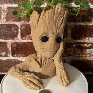 May include: A brown ceramic planter shaped like the character Groot from the Guardians of the Galaxy movie. The planter has a smiling face and is holding a plant with green leaves.