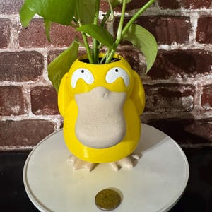 Op de afbeelding: Een gele Psyduck-vormige plantenpot met witte accenten, met een groene plant. De pot heeft een getextureerde snavel en staat op een wit oppervlak. Er ligt een munt voor de pot. De achtergrond is een bakstenen muur.