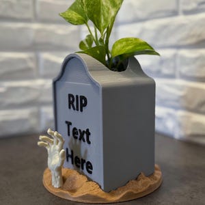May include: A gray tombstone-shaped planter with the text "RIP Text Here" and a zombie hand emerging from the base. The planter holds a green leafy plant. The base is a light brown color.