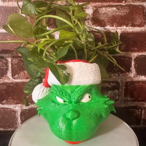 May include: A green Grinch planter with a white Santa hat, holding a green plant with long vines. The planter is sitting on a white surface.
