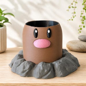 May include: A brown cylindrical planter with a smooth finish, featuring two black oval eyes with white highlights and a prominent pink oval nose. The planter is set within a textured, dark gray base that resembles a rocky, uneven ground surface.
