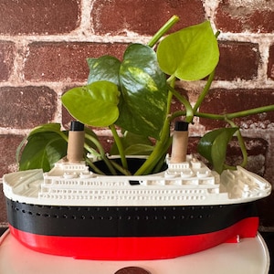 May include: A white, black, and red ship-shaped planter with a green plant growing out of it. The planter is sitting on a white surface.