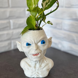 May include: A white, sculpted planter in the shape of a character's head with blue eyes and a grimacing expression. The planter holds a green leafy plant. The background is a white brick wall.