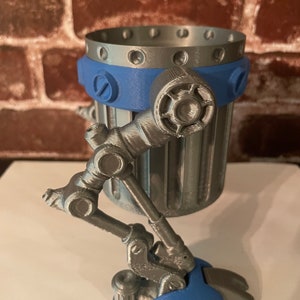 May include: A silver and blue 3D printed robot-shaped cup holder with a cylindrical body and a single leg. The leg is articulated and has a blue foot.