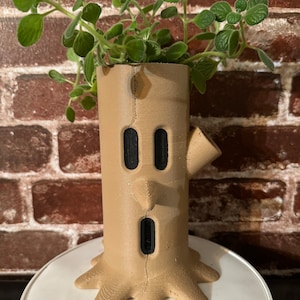 May include: A brown ceramic planter shaped like a tree trunk with a face. The planter has two black eyes, a black nose, and a black mouth. A green plant is growing out of the top of the planter.
