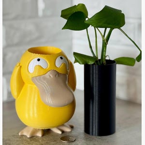 May include: A yellow Psyduck-shaped planter with a tan beak and feet, and white accents around the eyes. A black cylindrical planter holds a green plant with large leaves. A coin rests on the surface.