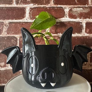 May include: A black ceramic planter shaped like a bat with white eyes and teeth. The planter has a small green plant growing inside.