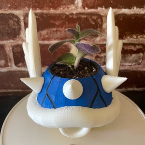 Könnte beinhalten: Ein blauer und weißer 3D-gedruckter Pflanztopf in Form eines Koopa Troopas aus der Super Mario Bros. Videospielserie. Der Topf hat eine kleine Pflanze darin.