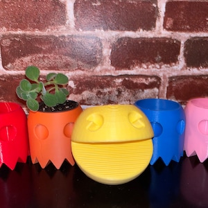 Puede incluir: Cinco macetas con forma de Pac-Man, una con una planta, en rojo, naranja, amarillo, azul y rosa.