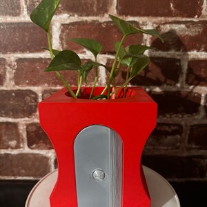 May include: A red pencil sharpener planter with a gray sharpening blade. A small green plant is growing inside the planter.