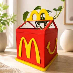 Peut inclure: Un pot de fleurs carré rouge en forme de boîte de repas rapide avec le logo jaune des arches dorées de McDonald's sur le devant et un graphique de sourire jaune sur le côté. Le dessus présente une poignée en arche jaune et une plante verte.
