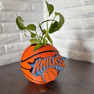 May include: A spherical orange basketball planter with black seams and a prominent blue, orange, and silver New York Knicks logo attached to the front. A green leafy plant grows from the top opening. A single copper penny sits at the base.