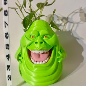 May include: A bright green, 3D-printed planter shaped like the Ghostbusters character, Slimer. The planter has a wide, open mouth with white teeth and a pink tongue. The eyes are yellow. The planter is holding a green plant with green and white leaves. The planter is approximately 5 inches tall.