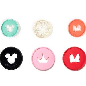 May include: Six round, colorful, plastic cookie cutters with different Disney character shapes: Mickey Mouse, Minnie Mouse, a crown, a bow, and two different ears. The cookie cutters are on a white background.