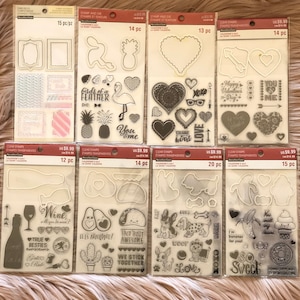 Puede incluir: Ocho láminas de sellos transparentes con varios diseños, incluyendo corazones, piñas, flamencos, botellas de vino, aguacates, tacos, perros y plátanos. Los sellos están empaquetados en plástico transparente con etiquetas marrones y blancas que indican el número de sellos por lámina y el precio.