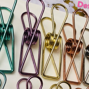 May include: Five metal binder clips in different colors: teal, purple, gold, copper, and silver. The clips are all in the shape of a rectangle with a spring in the middle. The word "Detail" is written in white letters on a black background in the top right corner of the image.