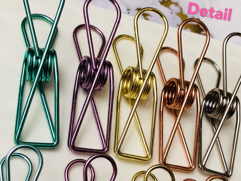 May include: Five metal binder clips in different colors: teal, purple, gold, copper, and silver. The clips are all in the shape of a rectangle with a spring in the middle. The word "Detail" is written in white letters on a black background in the top right corner of the image.