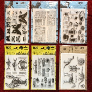 May include: Set of six clear transparent rubber stamps with various designs. The stamps are packaged in individual clear plastic sleeves with yellow and black borders. The stamps feature designs such as butterflies, coffee cups, a crab, a lobster, a fish, a dinosaur, a skeleton, and a spiral shell. The stamps are labelled with the brand name "Moodtape" and the product name "Transparent Stamps".