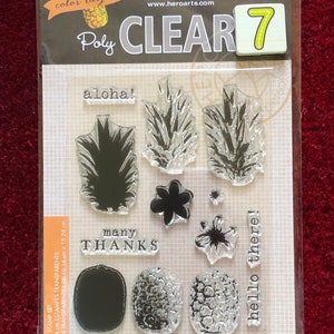 May include: A clear stamp set with seven stamps featuring pineapple designs, flowers, and the phrases "aloha!", "many thanks", "sending you warm wishes & sunny smiles", and "hello there!" The set is labeled "Poly Clear 7" and "Hero Arts".