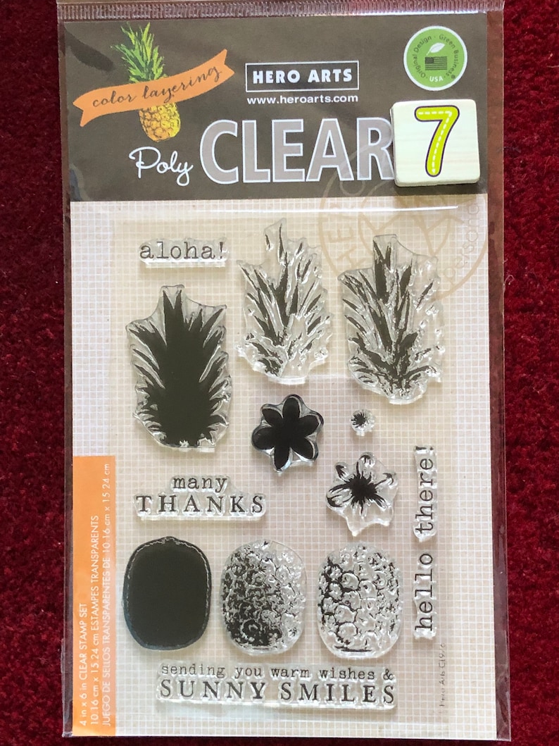 May include: A clear stamp set with seven stamps featuring pineapple designs, flowers, and the phrases "aloha!", "many thanks", "sending you warm wishes & sunny smiles", and "hello there!" The set is labeled "Poly Clear 7" and "Hero Arts".