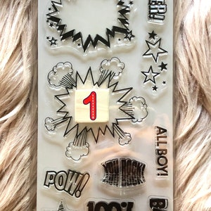 May include: Clear stamps with black ink featuring various comic book themed designs, including "POW!" "BAM!" "SUPER!" "YOU ROCK!" "100% ROCK STAR!" "ALL BOY!" and a star with a number 1 in a speech bubble.