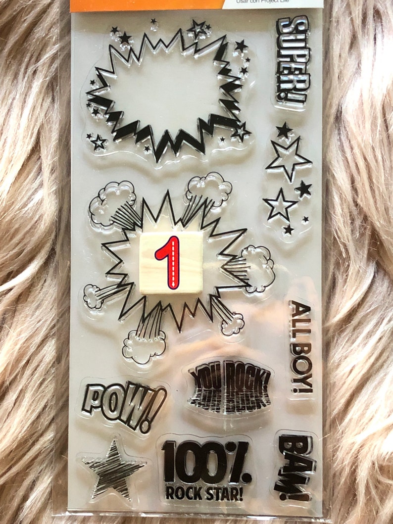 May include: Clear stamps with black ink featuring various comic book themed designs, including "POW!" "BAM!" "SUPER!" "YOU ROCK!" "100% ROCK STAR!" "ALL BOY!" and a star with a number 1 in a speech bubble.