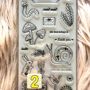 May include: A clear sheet of transparent stamps with various designs, including a snail, a bird, a mushroom, a flower, an owl, a bunny, and a caterpillar. The stamps are arranged in a grid pattern on a white background. The text "property of:", "snail mail", "this book belongs to:", "thank you", "hello", and "SPECIAL DELIVERY" are also included on the sheet.
