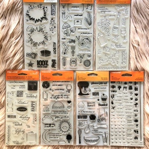 May include: A set of ten clear rubber stamps with black ink designs. The stamps are organized in two rows of five. The designs include various phrases, images, and numbers. The stamps are packaged in clear plastic sheets with orange and white borders.
