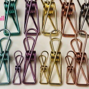 May include: Assortment of ten metal binder clips in various colors, including teal, purple, gold, copper, and silver. The clips are all the same size and shape, with a spring-loaded mechanism.