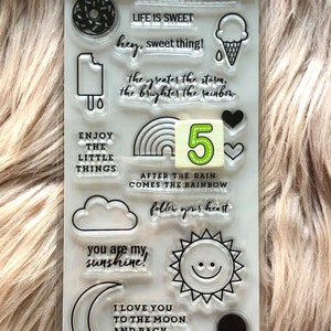 May include: A sheet of 20 clear stamps with various designs, including a donut, ice cream cone, rainbow, clouds, sun, moon, and text. The text includes phrases like "You melt my heart", "Life is sweet", "Enjoy the little things", and "After the rain comes the rainbow".