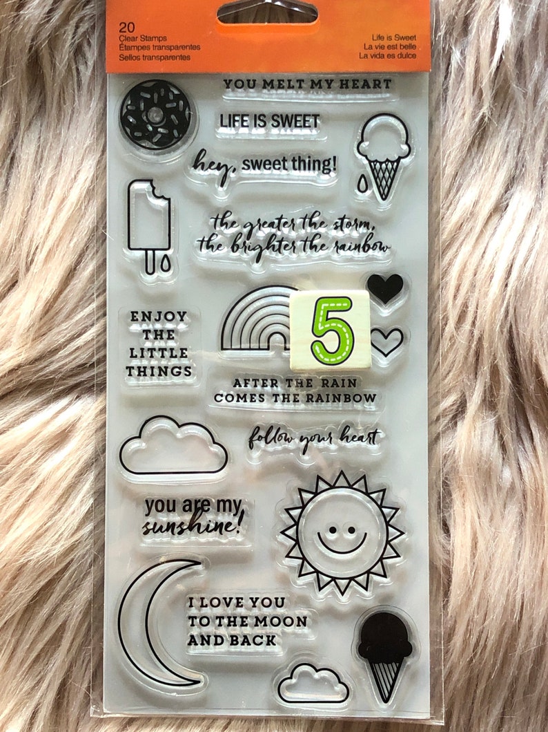 May include: A sheet of 20 clear stamps with various designs, including a donut, ice cream cone, rainbow, clouds, sun, moon, and text. The text includes phrases like "You melt my heart", "Life is sweet", "Enjoy the little things", and "After the rain comes the rainbow".