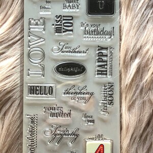 May include: A clear sheet of rubber stamps with various phrases and designs, including "LOVE", "just because", "sweet baby", "thinking of you", "you're invited", "congratulations", "with sympathy", "you're the best", "happy anniversary", "it's your birthday", "thank you", "hello", "feel better soon", and "love you". There is also a number "4" stamp.