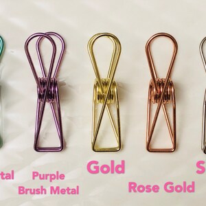 May include: Five metal binder clips in different colors: green, purple, gold, rose gold, and silver. The clips are all the same size and shape, and they are arranged in a row from left to right.