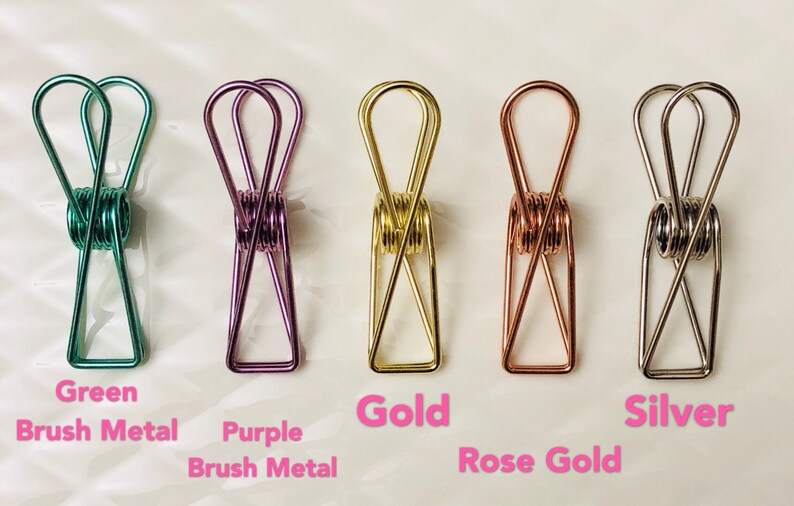 May include: Five metal binder clips in different colors: green, purple, gold, rose gold, and silver. The clips are all the same size and shape, and they are arranged in a row from left to right.