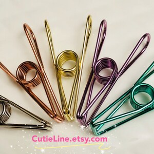 May include: Six metal binder clips in various colors, including copper, gold, purple, and green. The clips are arranged in a circle on a white background. CutieLine.etsy.com is printed at the bottom of the image.