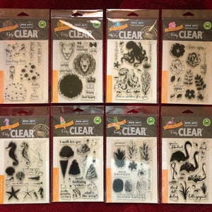 May include: Eight clear stamp sheets with various designs, including a lion, an octopus, flowers, a pineapple, a flamingo, and a seahorse. The stamp sheets are packaged in clear plastic and have a white background with black text that reads "Hero Arts Pretty Clear Stamps".