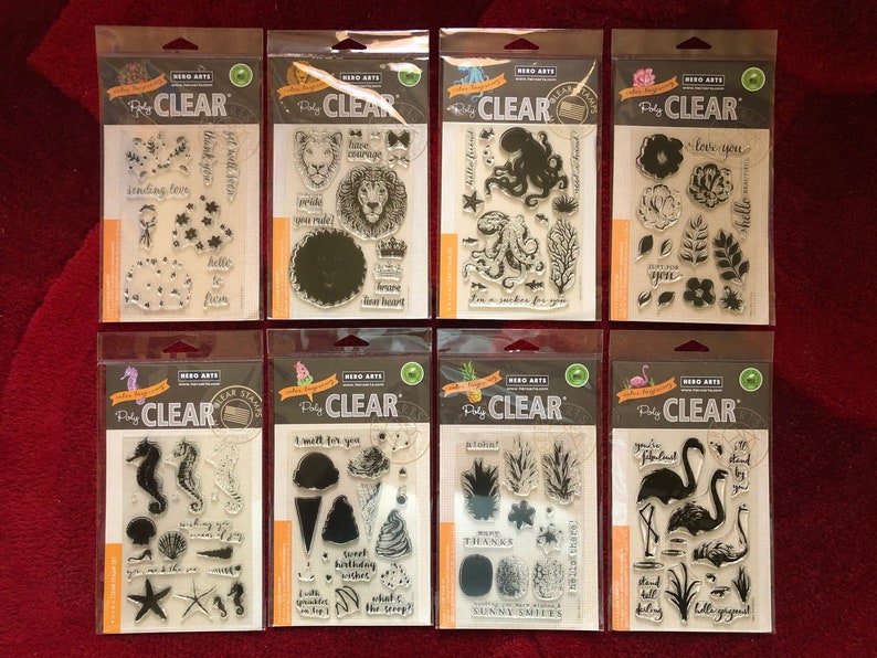 May include: Eight clear stamp sheets with various designs, including a lion, an octopus, flowers, a pineapple, a flamingo, and a seahorse. The stamp sheets are packaged in clear plastic and have a white background with black text that reads "Hero Arts Pretty Clear Stamps".