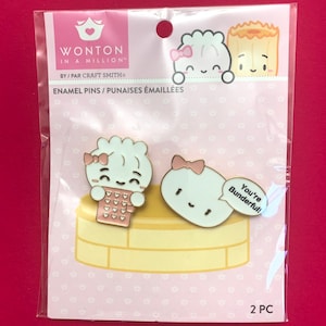 Puede incluir: Dos pines de esmalte con dibujos animados de albóndigas con lazos rosas. Una albóndiga sostiene un recipiente rosa para llevar con corazones y la otra albóndiga tiene un globo de diálogo que dice "You're Bunderful!" Los pines están empaquetados en una tarjeta rosa y blanca con el texto "Wonton in a Million™ by/Par Craft Smith Enamel Pins/Punaises Émaillées 2 PC".