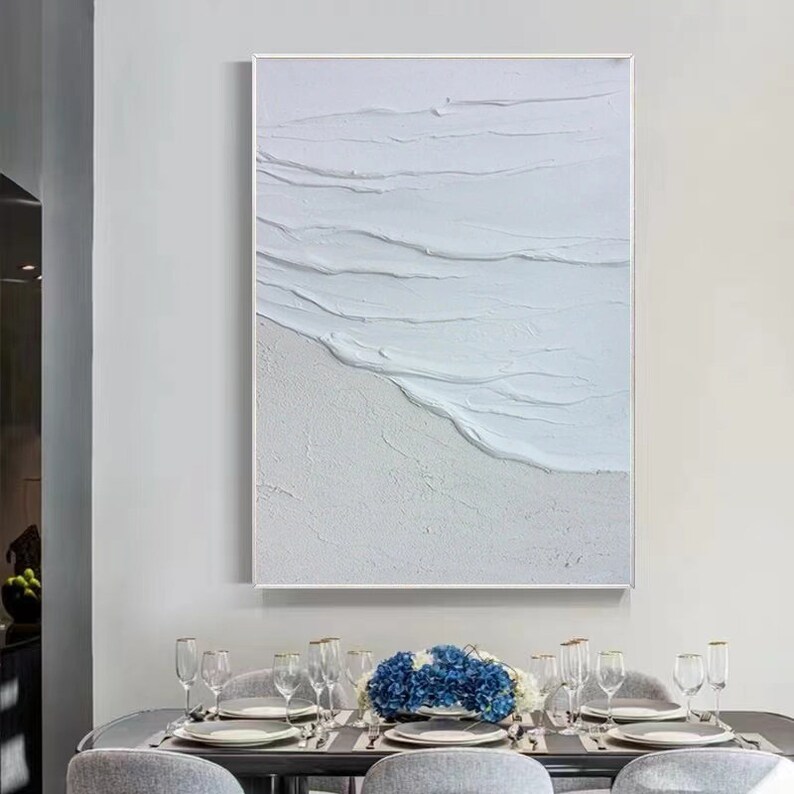 May include: White abstract painting of waves crashing on a beach. The painting is framed in a white frame and hangs on a white wall above a dining table.