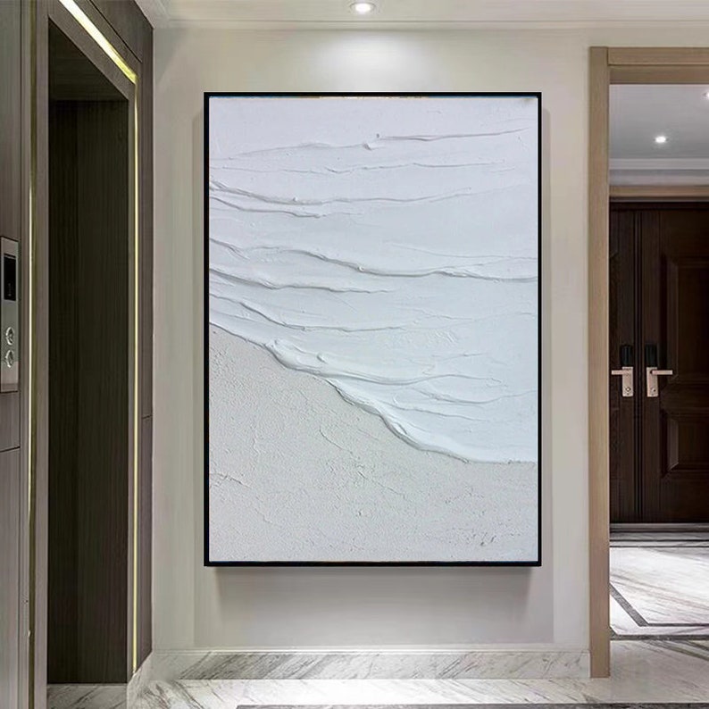 May include: A white abstract painting with textured brushstrokes that resemble waves. The painting is framed in a black frame and is hanging on a white wall.