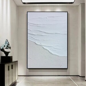 May include: White abstract painting with textured brushstrokes resembling waves on a beach. The painting is framed in a black frame and hangs on a white wall.
