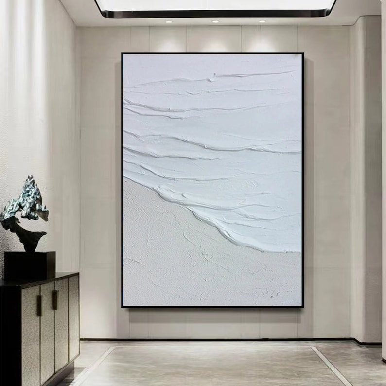 May include: White abstract painting with textured brushstrokes resembling waves on a beach. The painting is framed in a black frame and hangs on a white wall.