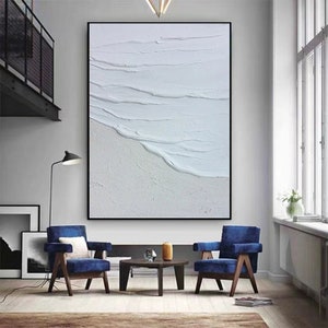 May include: A white abstract painting with textured brushstrokes depicting waves crashing on a beach. The painting is framed in black and hangs on a white wall in a living room setting.