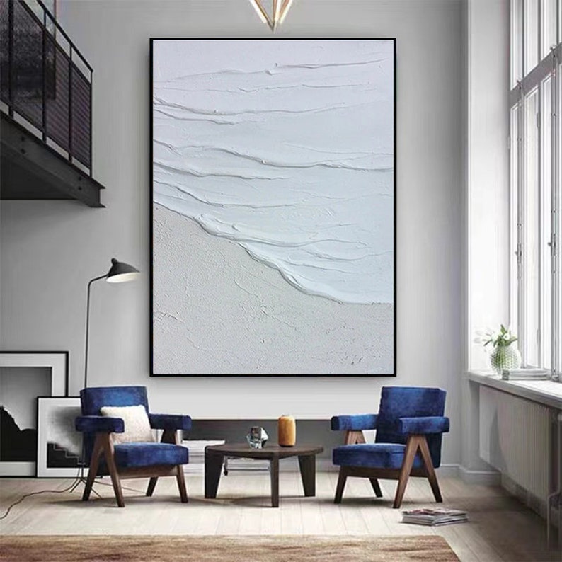 May include: A white abstract painting with textured brushstrokes depicting waves crashing on a beach. The painting is framed in black and hangs on a white wall in a living room setting.