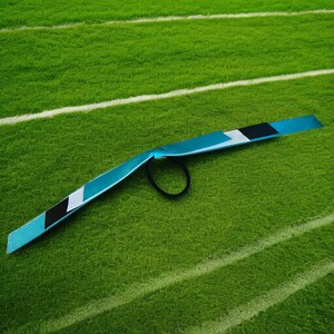 May include: A teal hair accessory with a black elastic band. The accessory has a rectangular shape with a black, white, and grey stripe design. The item is lying on a green surface with white lines.
