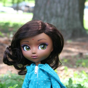 Könnte beinhalten: Eine Puppe mit langen, dunkelbraunen Haaren, grünen Augen und einem blau-weiß gemusterten Shirt. Die Puppe hat einen kleinen roten Punkt auf der Stirn.